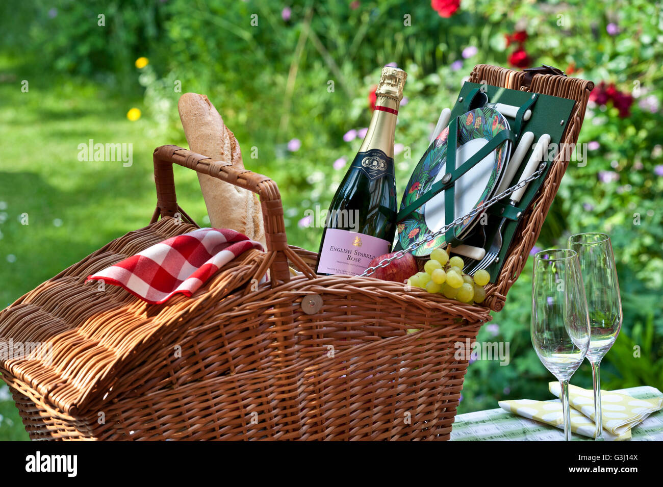 Englische Sekt-Rosé-Flasche und Picknickkorb mit Baguette und Trauben in sonniger, blumiger englischer Gartenlage Stockfoto