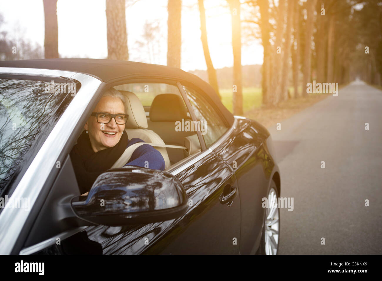 Frau im Auto fahren auf Baum gesäumten Straße Blick aus Fenster lächelnd Stockfoto