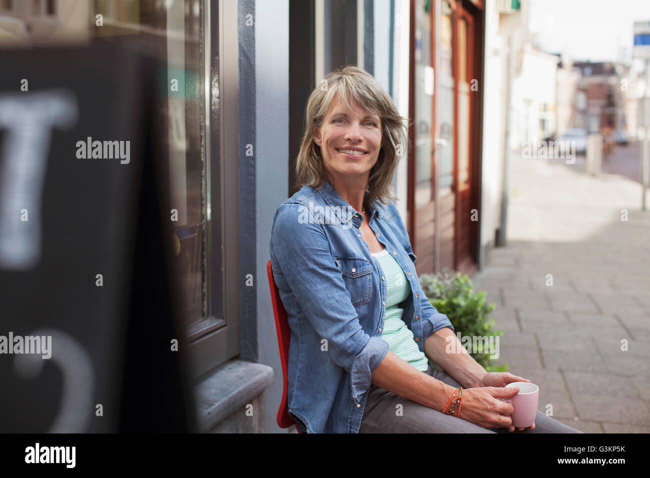 Frau sitzt auf dem Stuhl vor Geschäft halten Kaffeetasse Blick auf die Kamera zu Lächeln Stockfoto