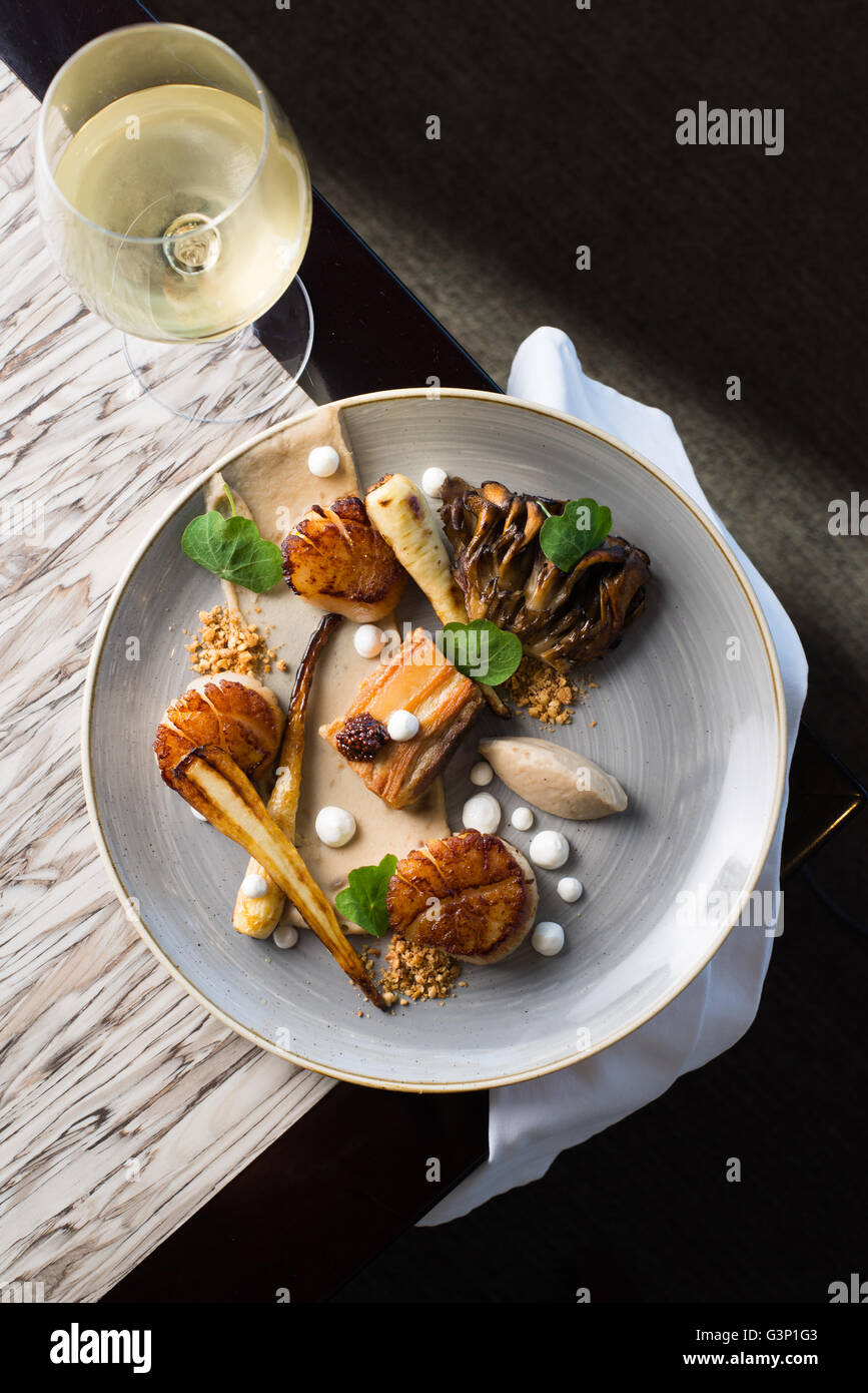 Draufsicht einer Jakobsmuschel-Medley-Platte mit einem Glas Weißwein in einem feinen Restaurant Ambiente. Stockfoto