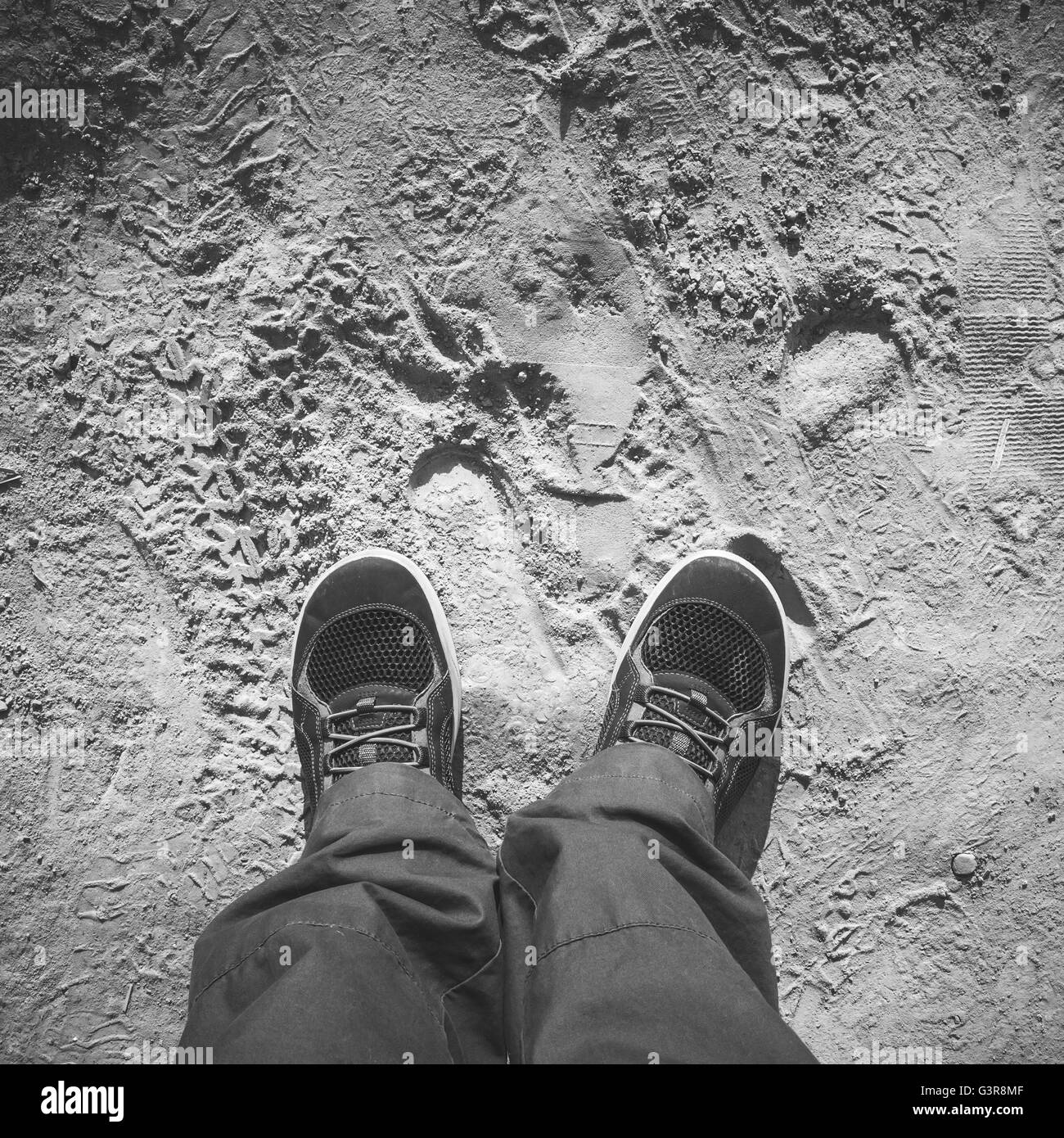 Männliche Füße in sportlichen Schuhen stehen auf schmutzigen staubigen Straße, Egoperspektive, eckig schwarz / weiß Foto Stockfoto