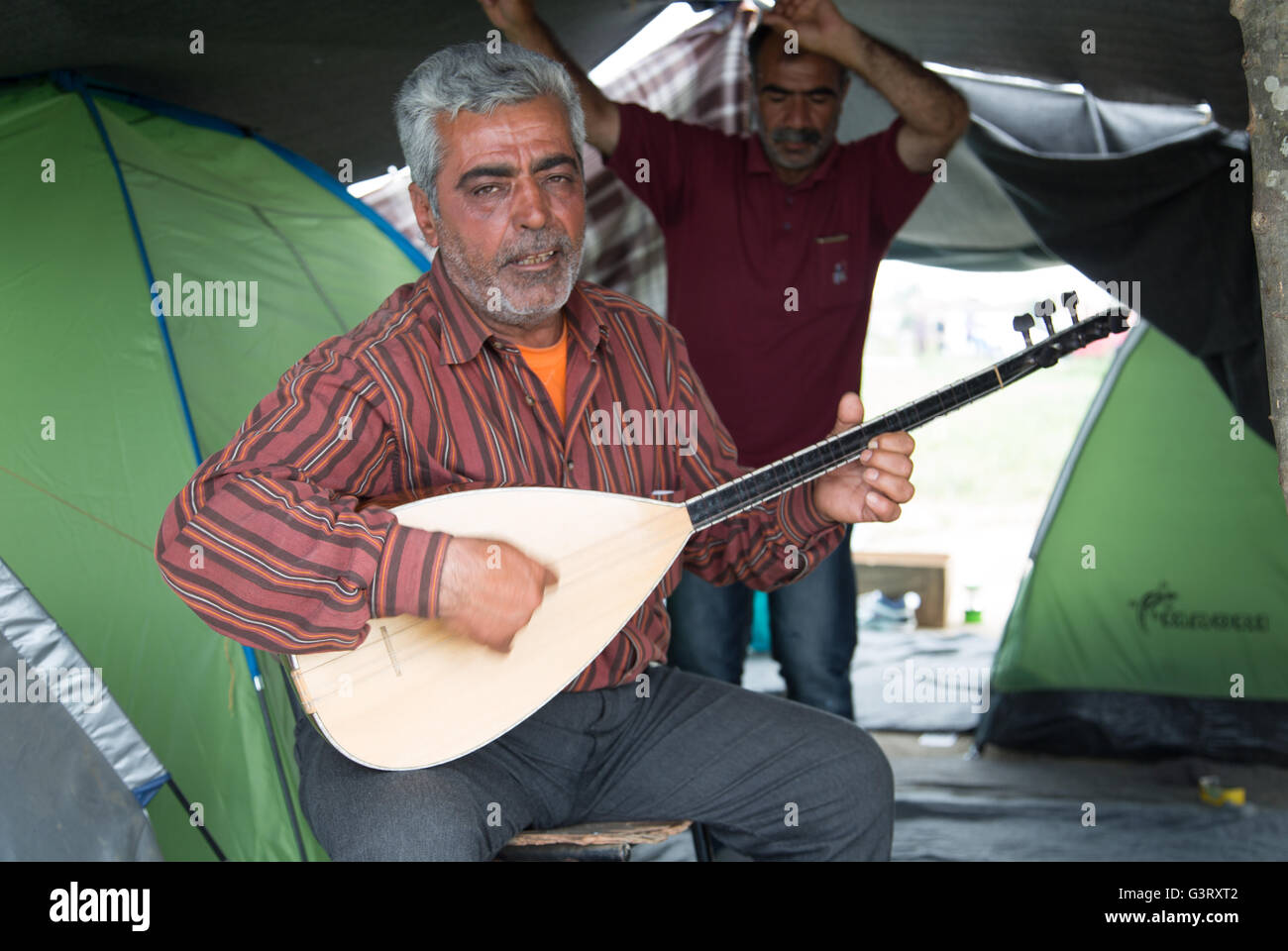Ein kurdischer syrischen Mann spielt ein traditionelles Instrument, Oud, während sein Freund Tänze im Hintergrund im Flüchtlingslager Idomeni Stockfoto