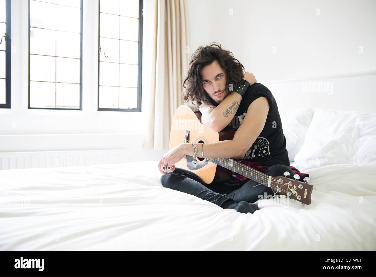 Ein cooler junger Mann sitzt auf einem Bett mit einer Gitarre. Stockfoto
