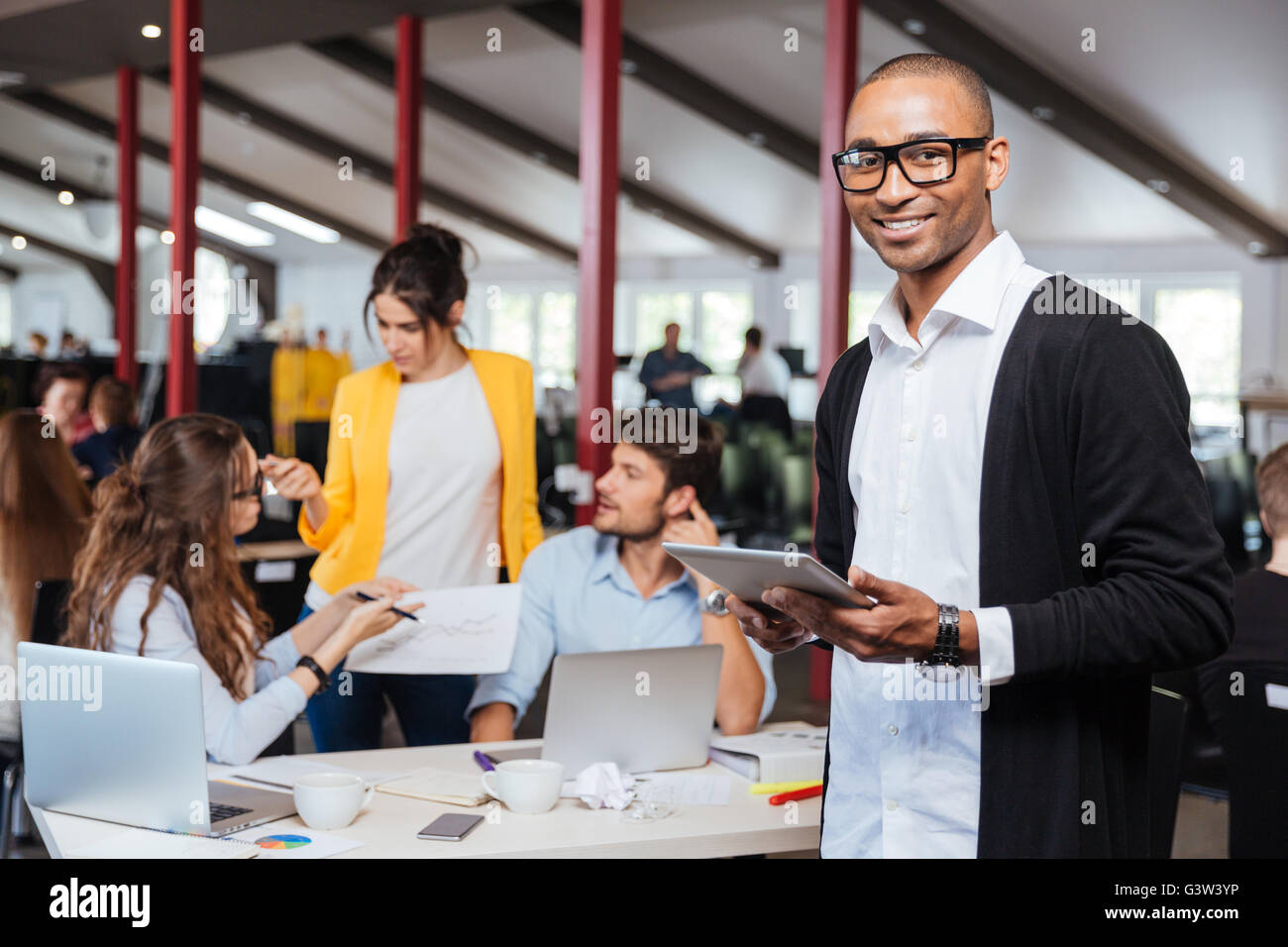 Fröhliche afrikanische Jungunternehmer mit Tablet im Büro mit Kollegen im Hintergrund Stockfoto