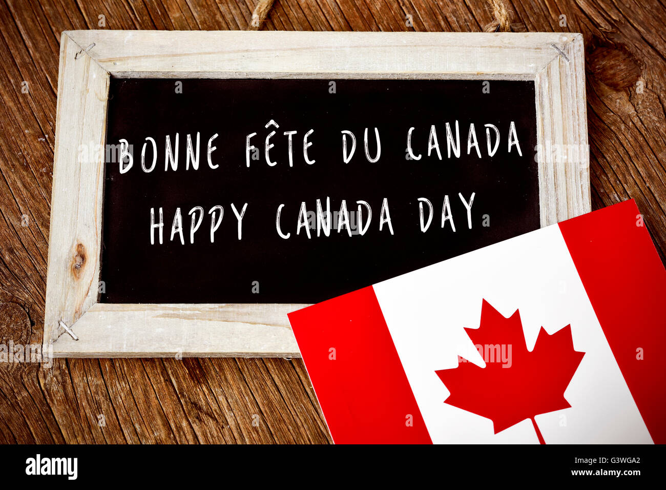 der Text Happy Canada Day geschrieben in Französisch und Englisch in einer Tafel und eine Fahne von Kanada, auf einer rustikalen Holzoberfläche Stockfoto