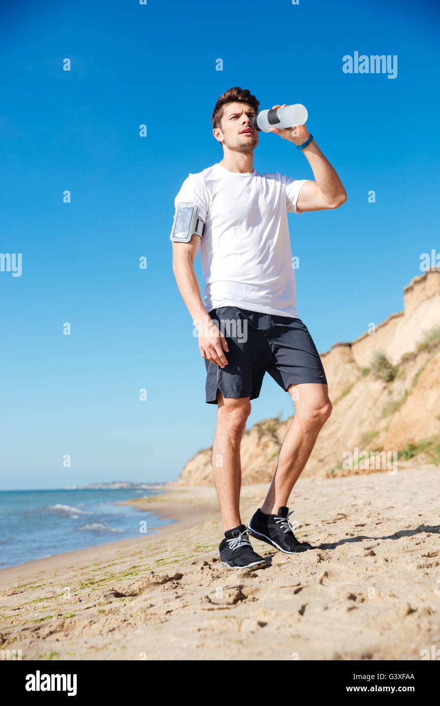 Hübscher Junge Sportler stehen und Trinkwasser am Strand Stockfoto