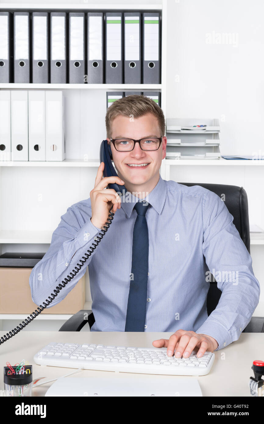 Lächelnde Jungunternehmer mit Brille ist telefonieren und tippen auf der Computertastatur beim Sitzen am Schreibtisch im Büro. A Stockfoto