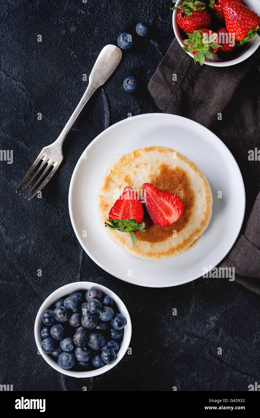 Pfannkuchen mit Erdbeeren und Heidelbeeren in weißer Keramik Teller und Schalen mit frischen Beeren auf Textile Serviette über schwarz tex Stockfoto