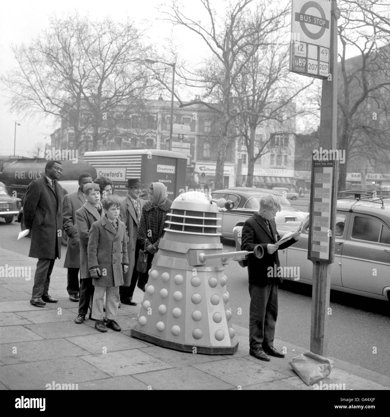 Ein junger anwartender Passagier liest seine Zeitung, ohne den Dalek aus der BBC-Fernsehsendung „Dr. Who“ zu kennen, in der Schlange hinter ihm. Stockfoto