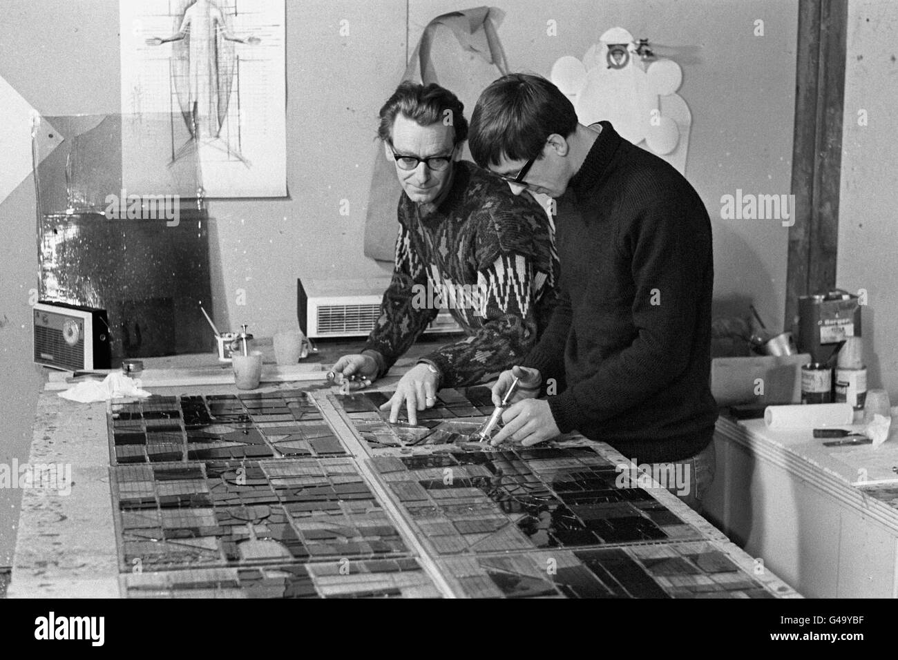 John Hayward (l) und Stephen Taylor verwenden hypodermische Spritzen, um den Klebstoff aufzutragen, wenn sie Glasmalerei an Plattenglas für eine der Platten einer riesigen Laterne kleben, die sie für Blackburn Cathedral, Lancashire, machen. Sie arbeiten in einem umgebauten Stall bei Mr Hayward's Haus in Prickloves Farmhouse, Bletchingley, Surrey. John, ursprünglich ein Möbeldesigner, aber jetzt konzentriert sich auf Glasmalerei, entworfen die Laterne, die fast 1,000 Quadratmeter Glasmalerei auf einem abstrakten Thema basiert enthalten wird. In der Wand im Hintergrund ist eine Planzeichnung der 30ft Stahlfigur von Christus, Stockfoto