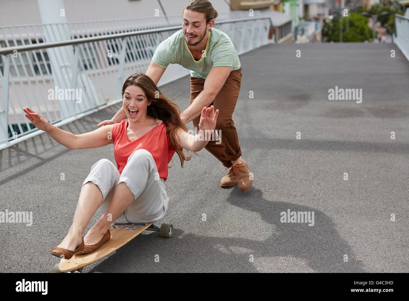 -MODELL VERÖFFENTLICHT. Junge Frau sitzt auf Skateboard mit Mann schieben. Stockfoto