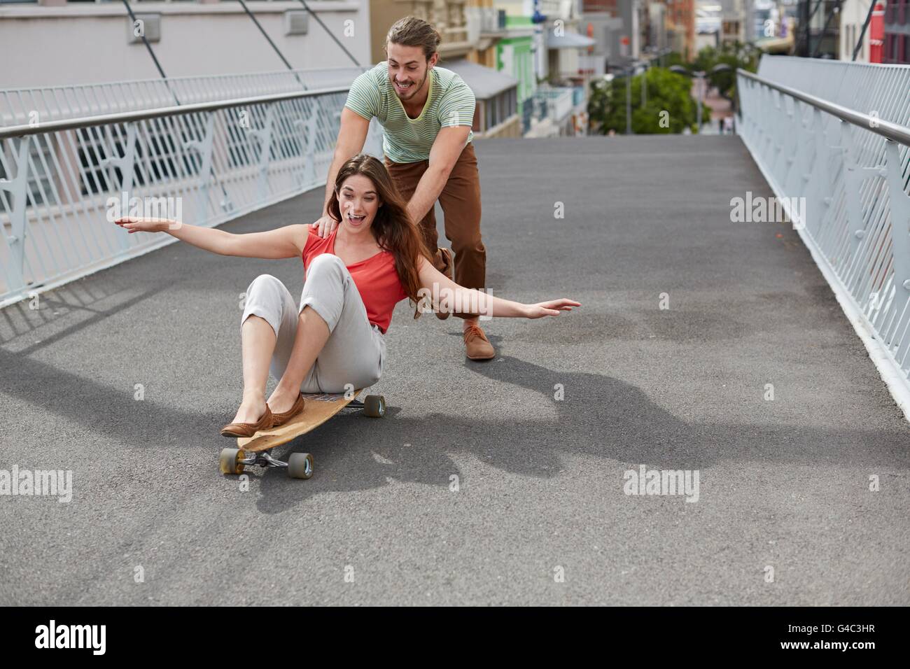 -MODELL VERÖFFENTLICHT. Junge Frau sitzt auf Skateboard mit Mann schieben. Stockfoto