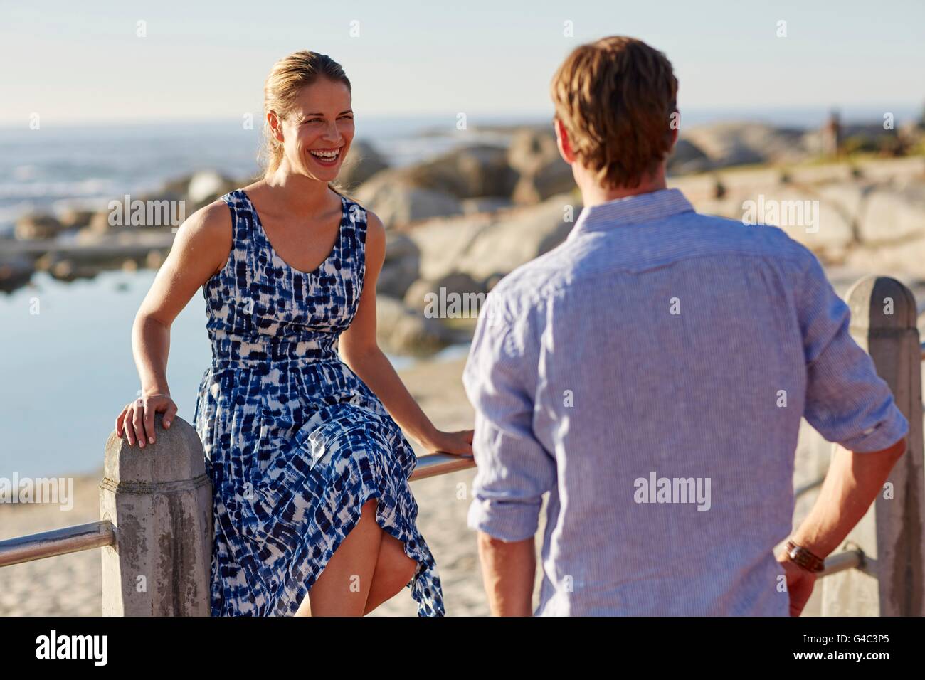 -MODELL VERÖFFENTLICHT. Frau sitzt am Geländer vom Strand lachen, Mann beobachtet. Stockfoto