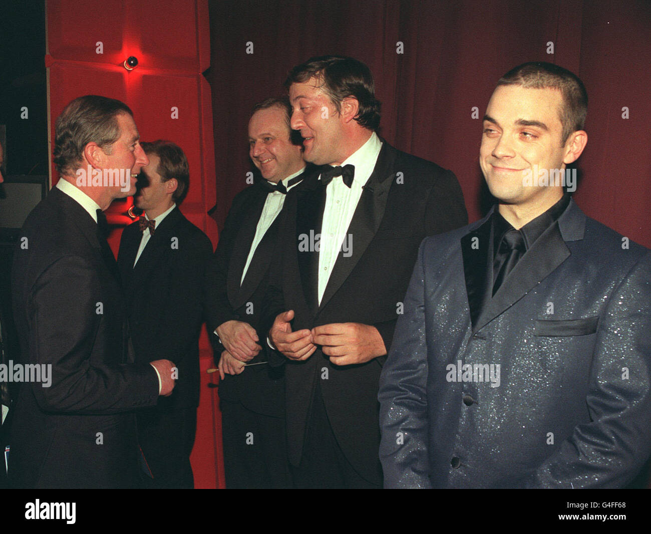 Der Prinz von Wales spricht hinter der Bühne mit Stephen Fry, während Robbie Williams (rechts) nach der Prince's Trust Comedy Gala im Lyceum Theatre in London aufschaut. Stockfoto