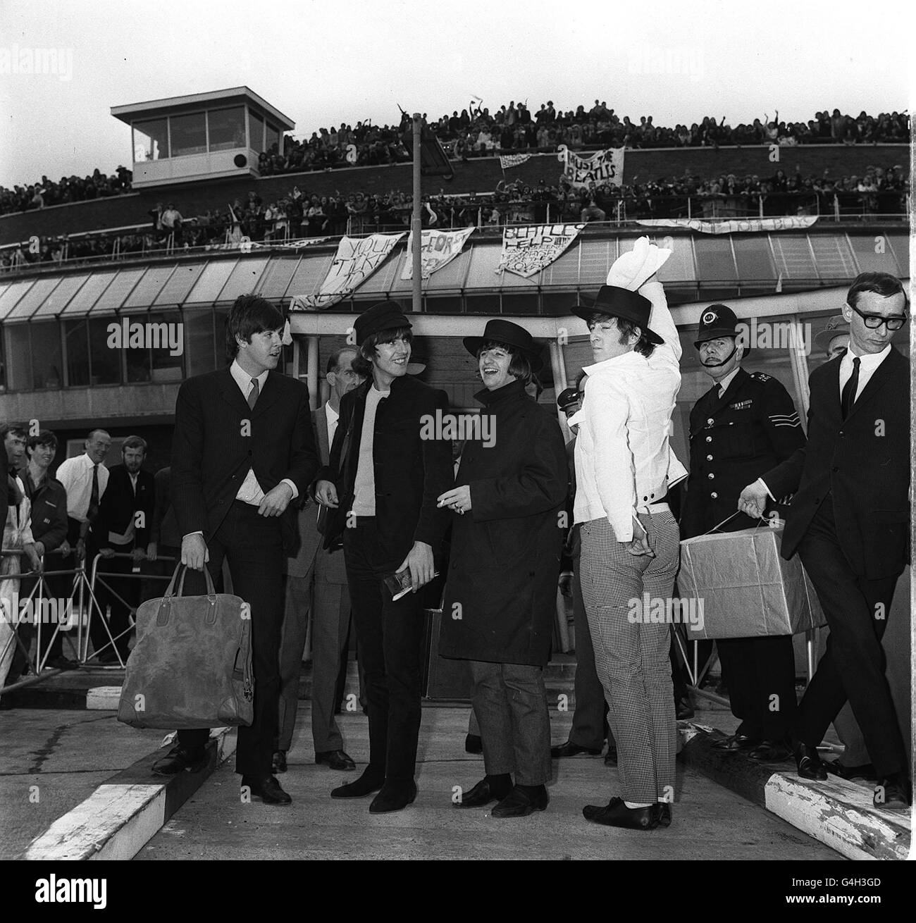 John Lennon, in einem Souvenir-Hut, schlägt eine spanische Pose für die Hunderte von Fans, die die Beatles - Paul McCartney, George Harrison & Ringo Starr auf dem Londoner Flughafen bei ihrer Rückkehr aus Spanien - schreiend begrüßen. 4/7/65 117349-1 ghgal Stockfoto