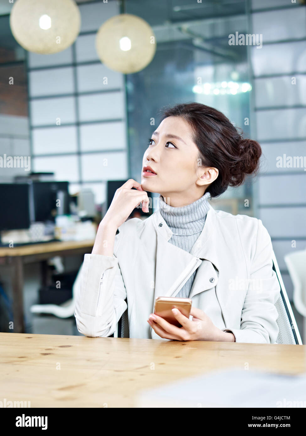 junge asiatische Geschäftsfrau im Büro nach oben denken mit dem Handy in der hand sitzen. Stockfoto