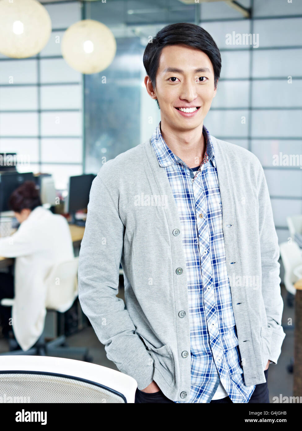 zuversichtlich asiatischen Jungunternehmer im Büro Blick in die Kamera stehen Hände in den Taschen lächelnd. Stockfoto
