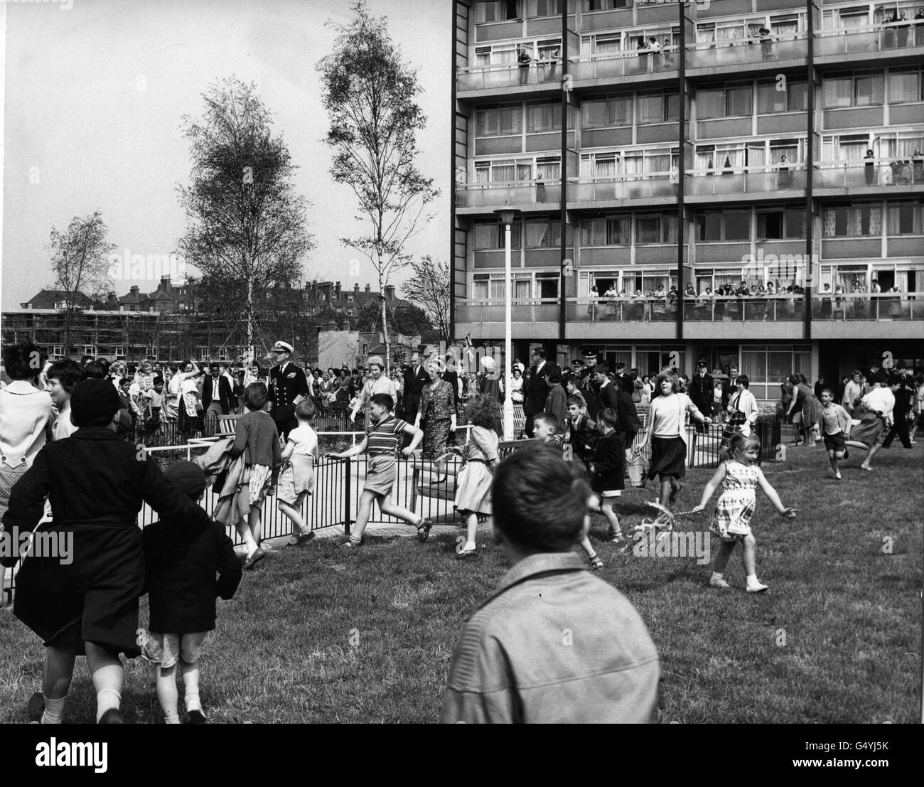 *eingescannte Low-Res from Print* Kinder laufen aufgeregt über das Gras, als Queen Elizabeth II (Mitte, leichter Mantel) und der Duke of Edinburgh in der Nähe des Wickham House (Hintergrund) auf dem Stiffford Estate, Stepney, im Londoner East End spazieren. Stockfoto