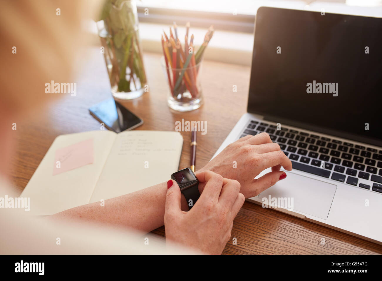 Schuss von Frau mit Smartwatch hautnah. Fokus auf weibliche Hände und Smartwatch mit Laptop und Tagebuch auf Tisch. Stockfoto