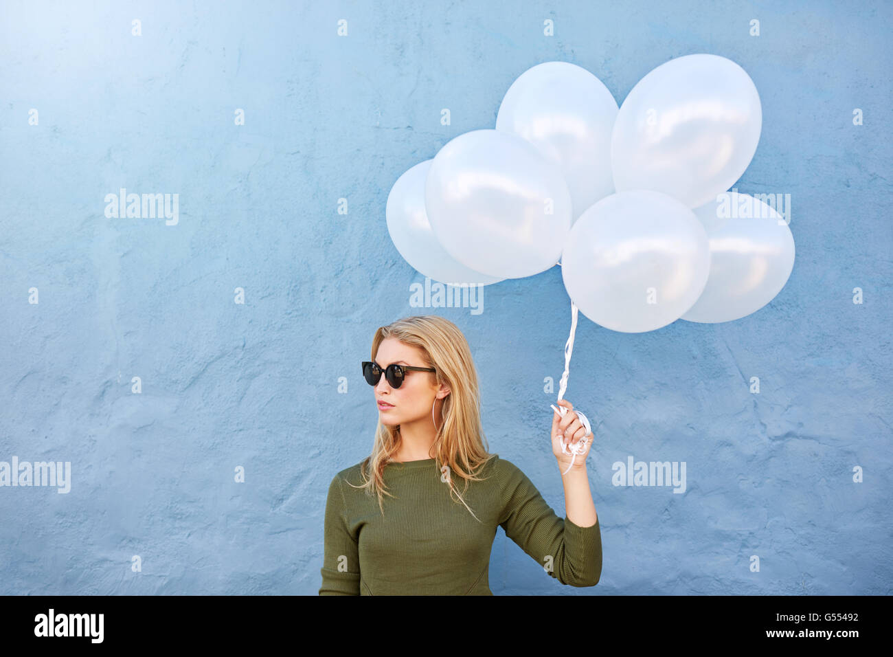 Schuss von trendige junge Frau mit Sonnenbrille Ballons halten und wegschauen gegen blaue Wand. Stockfoto