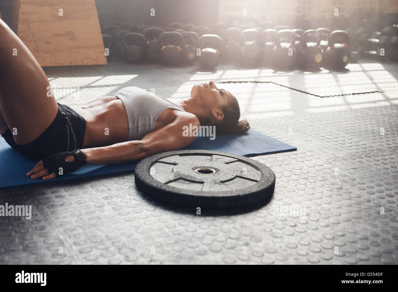 Fitness-Frau, die sich nach ihrem Training im Fitnessstudio entspannt. Schwere Platte auf dem Boden mit muskulöser Frau, die auf der Trainingsmatte liegt. Stockfoto