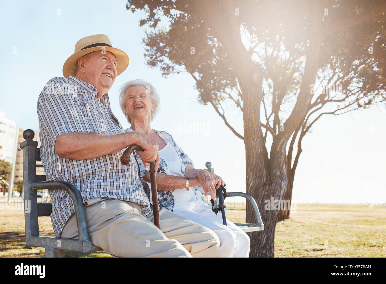 Glücklich älterer Mann und Frau sitzen auf einer Bank mit Spazierstock an einem Sommertag. Entspannte älteres Paar sitzt auf einer Parkbank Stockfoto