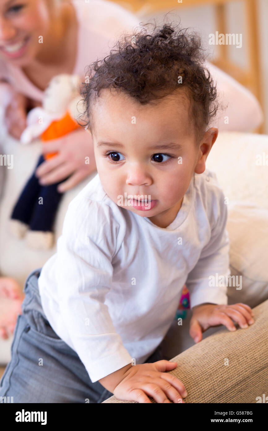 Baby Junge stehend und stützte sich auf der Rückseite des Sofas, mit seiner Mutter hinter ihm beobachtete ihn Stockfoto