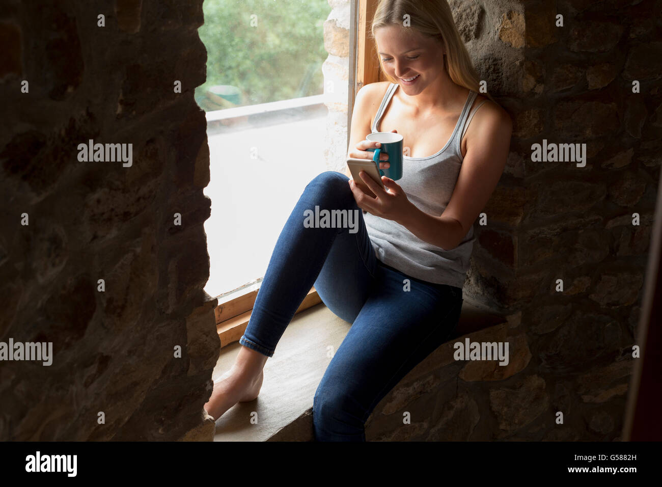 Frau auf ihrer Fensterbank sitzen, während eine Smartphone benutzen und genießen Sie ein heißes Getränk. Stockfoto