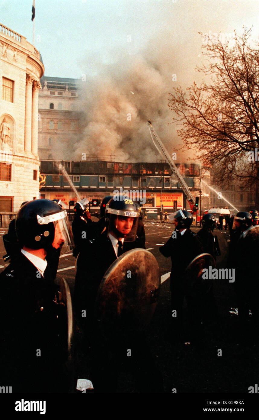 Polizeibeamte in Bereitschaftskleidung stehen am Trafalgar Square in London, nachdem sich ein Protest gegen die sogenannte Poll Tax zu einem Aufstand entwickelt hatte. Ein Gebäude am Strand ist im Hintergrund brennend zu sehen. Stockfoto