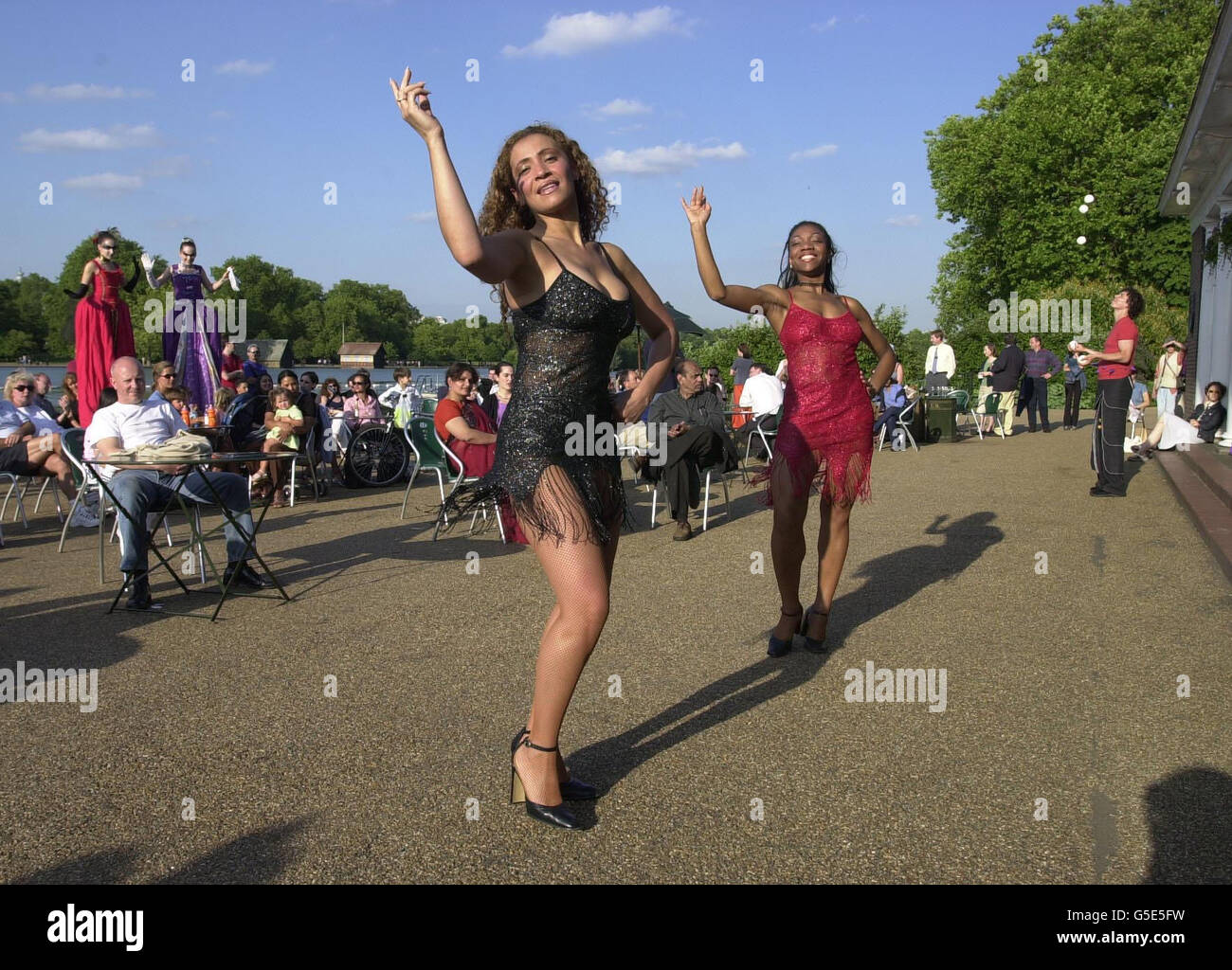 Kolumbianische Salsa-Tänzer togue de Queda (vorne), Stelzenläufer die Aristokratischen Damen (hinten links) und ein Jongleur amüsieren Café-Besucher am Serpentine Lake, im Londoner Hyde Park, bei der Eröffnung des Royal Parks Sommer-Entertainment-Programm. Stockfoto