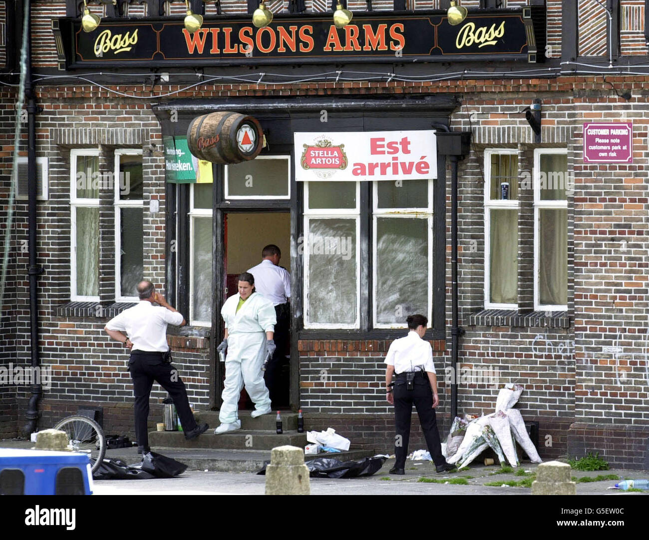 Polizeibeamte suchen im Wilsons Arms Pub in Leeds nach, wo ein Familienvater niedergeschossen wurde, während Kinder Party-Spiele spielten. Der frühere Profi-Rugby-League-Star David Nelson, 38, wurde am 22/07/01 im Pub in Seacroft in den Kopf geschossen. *...auf der rechten Seite sind Floral Tributes an Herrn Nelson angehäuft. Stockfoto