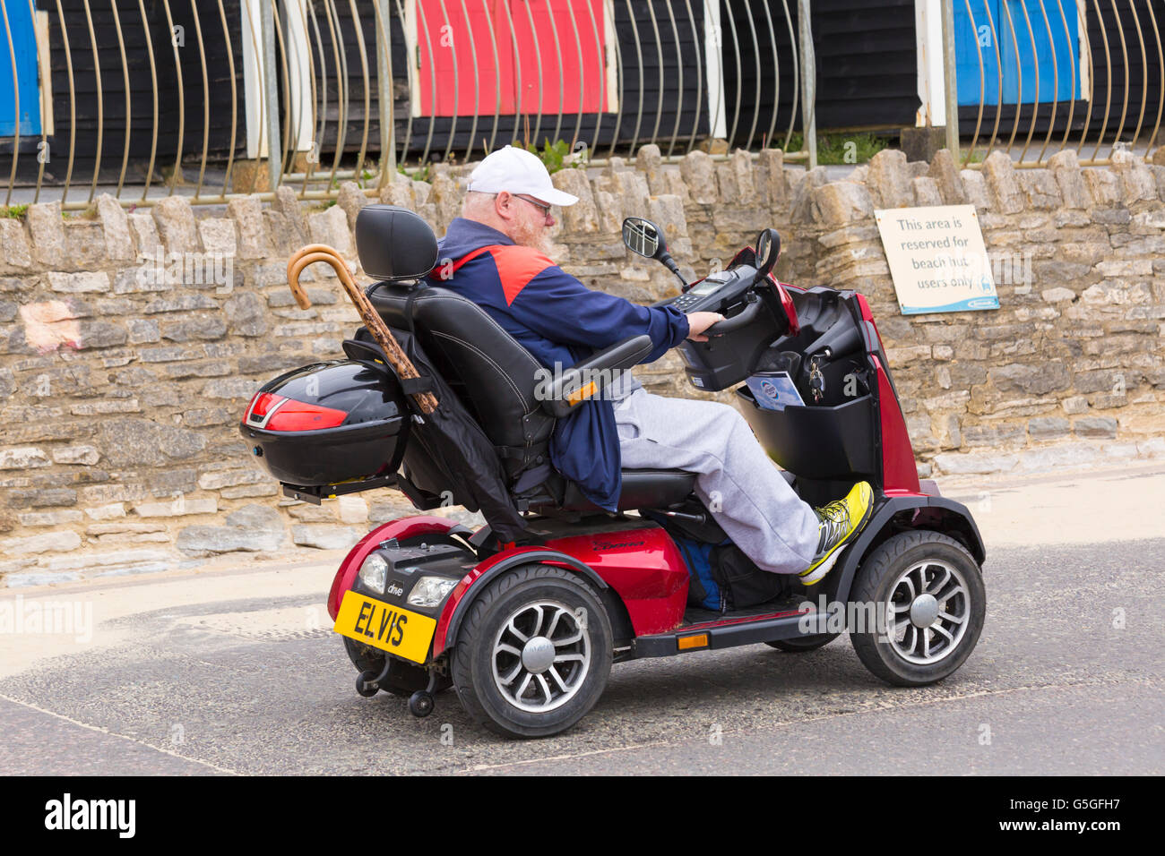 Älterer Mann auf der Mobilität Roller Elvis Fahrt vorbei an Reihe von Strandhütten auf Promenade in Bournemouth im Juni Stockfoto