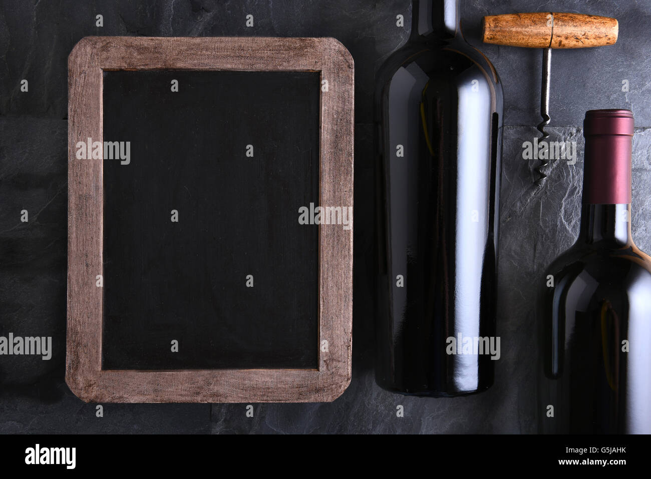 Draufsicht auf eine leere Tafel für eine Weinkarte oder ein Menü mit zwei Flaschen Wein und alte Korkenzieher. Streiflicht auf Schiefer Tisch. Stockfoto