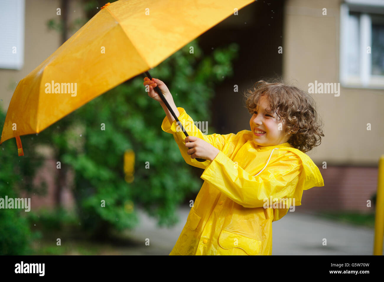 Der junge versucht 8-9 Jahre in einem hellen gelben Regenmantel, einen Regenschirm aus Wind Fehler halten. Er hält einen Regenschirm mit beiden Händen. Ch Stockfoto