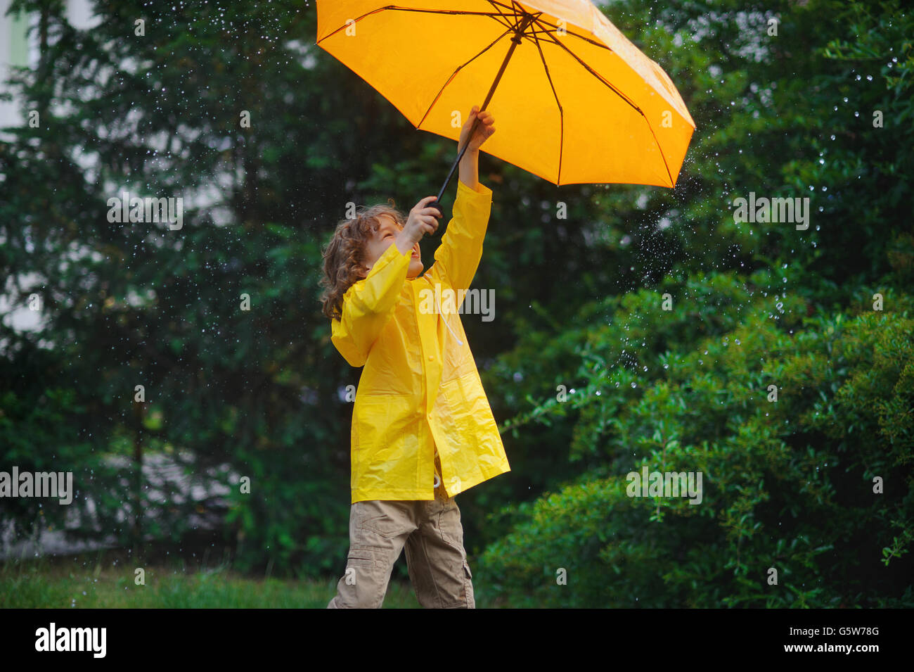 Kerlchen mit einem leuchtend gelben Schirm und in einem Regenmantel unter warmen Sommerregen. Er liebt den Regen und freut sich. Er fröhlich spielen Stockfoto