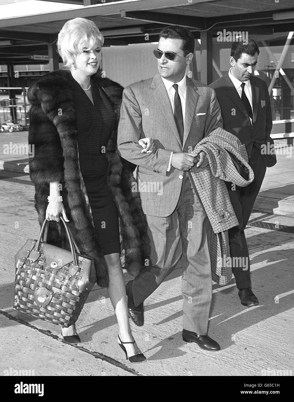 Die Hollywood-Schauspielerin Jayne Mansfield lächelt, als sie Arm in Arm mit ihrem Freund Enrico Bomba, einem italienischen Filmvorführer, am Flughafen London geht. Stockfoto