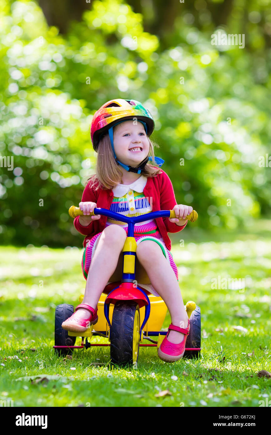 Niedliche Mädchen reiten ihr Dreirad im sonnigen Sommerpark-Schutzhelm tragen. Kinder Fahrrad. Erstes Fahrrad für kleines Kind Stockfoto
