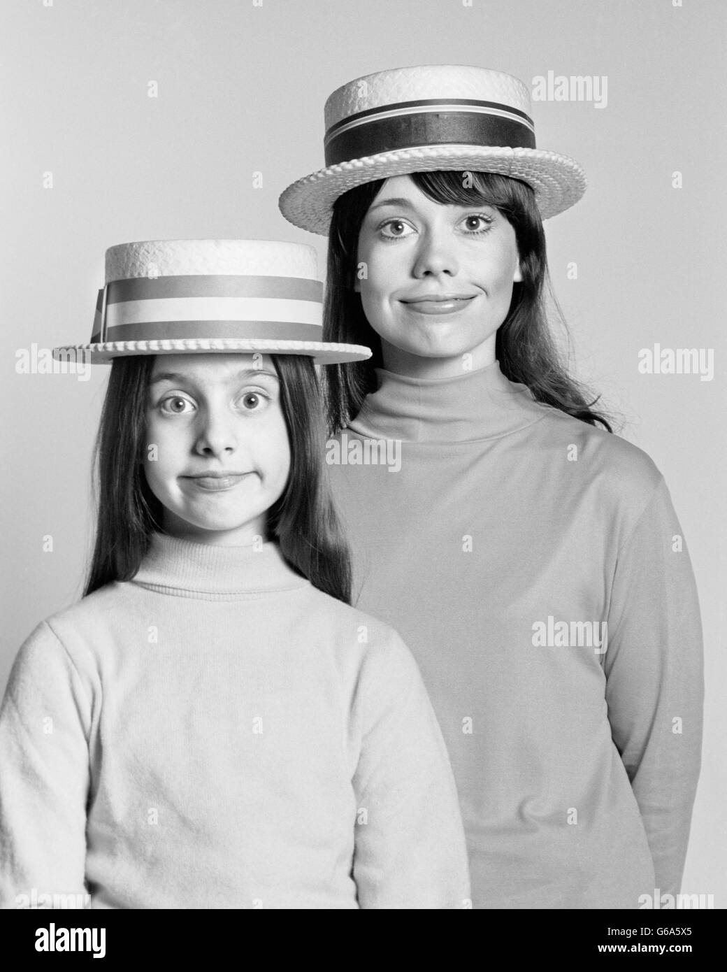 1970ER JAHRE MUTTER UND TOCHTER PORTRAIT AUSSEHEN GLEICHERMAßEN DUMMEN GESICHTSAUSDRUCK STROH BOOTSFAHRER HÜTE Stockfoto