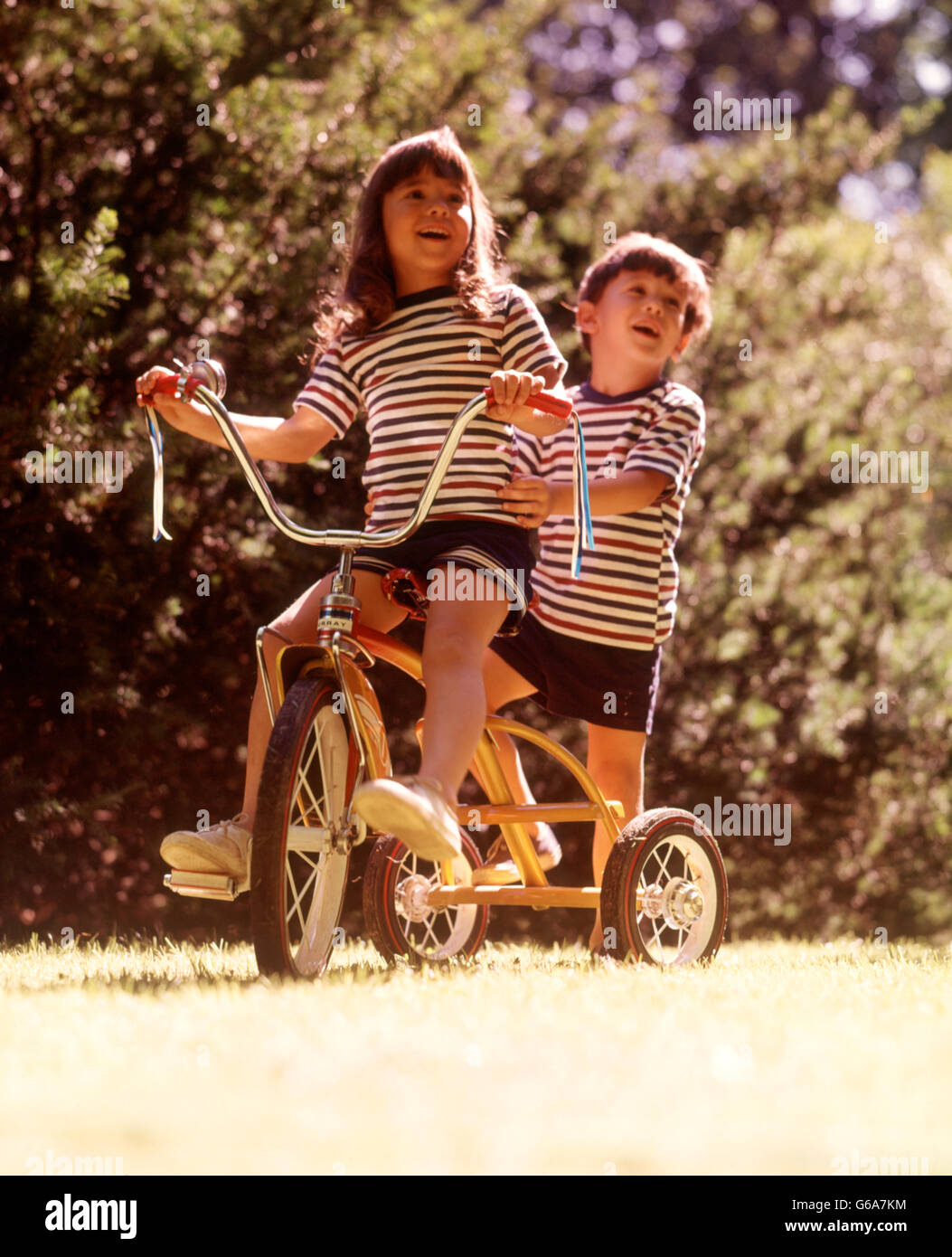 1970ER JAHREN GESTREIFT 2 KINDER T SHIRTS SHORTS FAHRRAD GELB DREIRAD FAHRRAD MÄDCHEN LENKUNG JUNGE STEHEND ZURÜCK Stockfoto