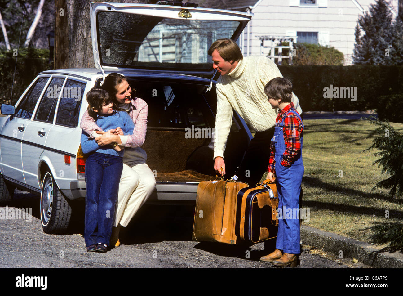 1980ER JAHRE VIERKÖPFIGE FAMILIE IMMER BEREIT FÜR DIE REISE-MUTTER UND TOCHTER SITZEN AUF AUTO HECKKLAPPE VATER UND SOHN TRAGEN KOFFER Stockfoto