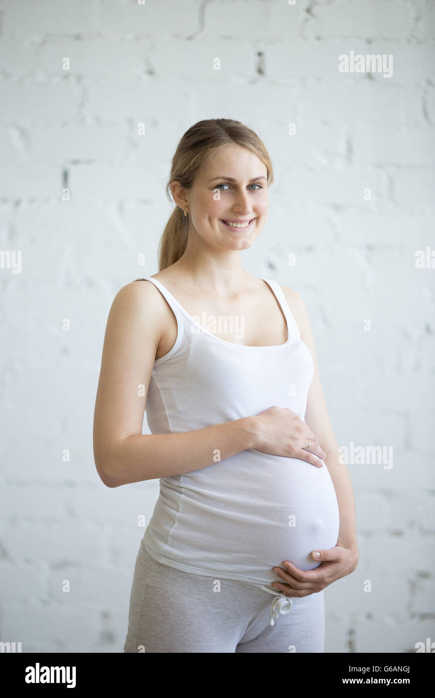 Gesunde Schwangerschaft Konzept. Porträt von schönen blonden junge schwangere Modell Blick in die Kamera, während Sie ihren Bauch streicheln Stockfoto