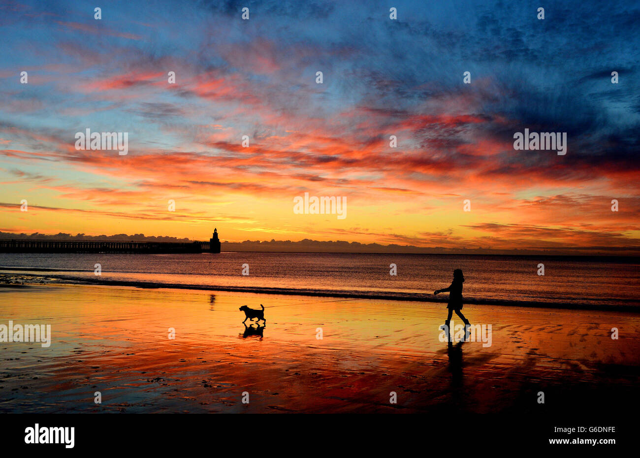 Eine Frau geht mit ihrem Hund bei Sonnenaufgang am Blyth Beach, Northumberland. Stockfoto