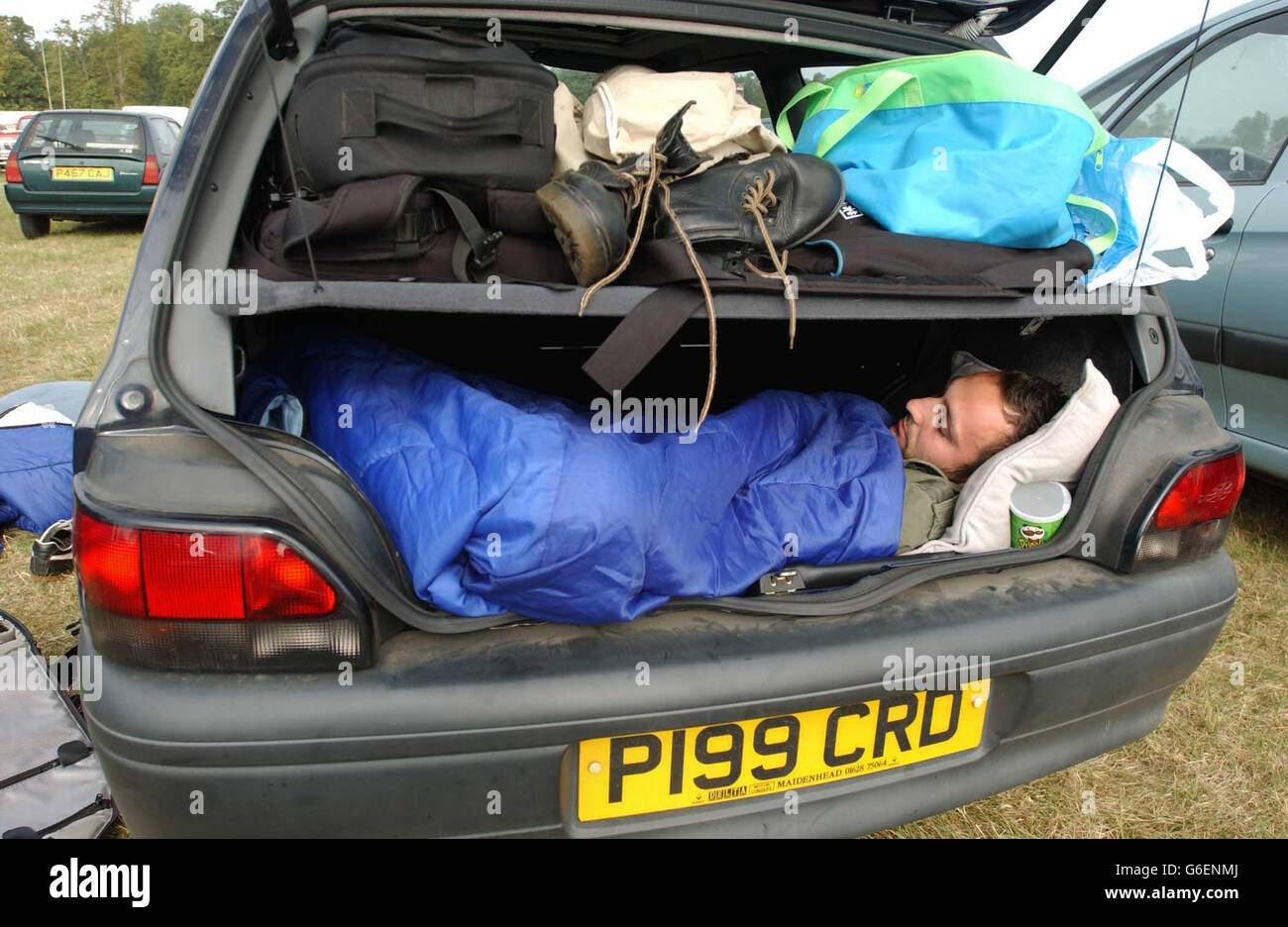 Ein Nachtschwärmer schläft im Kofferraum eines Autos beim V2003 Musikfestival im Hylands Park, Chelmsford, Essex. Tausende von Feiernden genossen am zweiten Tag des Festivals weiterhin die Sonne, mit Bands wie Inspiral Carpets, Morcheeba und David Gray, wobei die Red Hot Chili Peppers das Event zu Ende brachten. Stockfoto