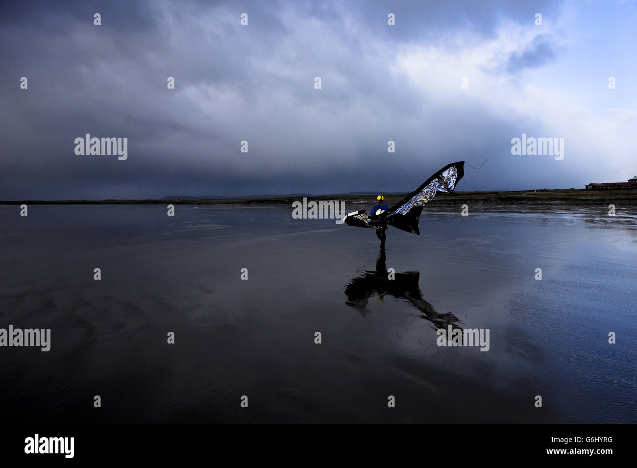 Ein Kitesurfer läuft am Strand bei starkem Wind und dramatischem stürmischen Wetter mit seinem Drachen an seinem Rücken in Rest Bay, Porthcawl, Wales geschnallt. Stockfoto