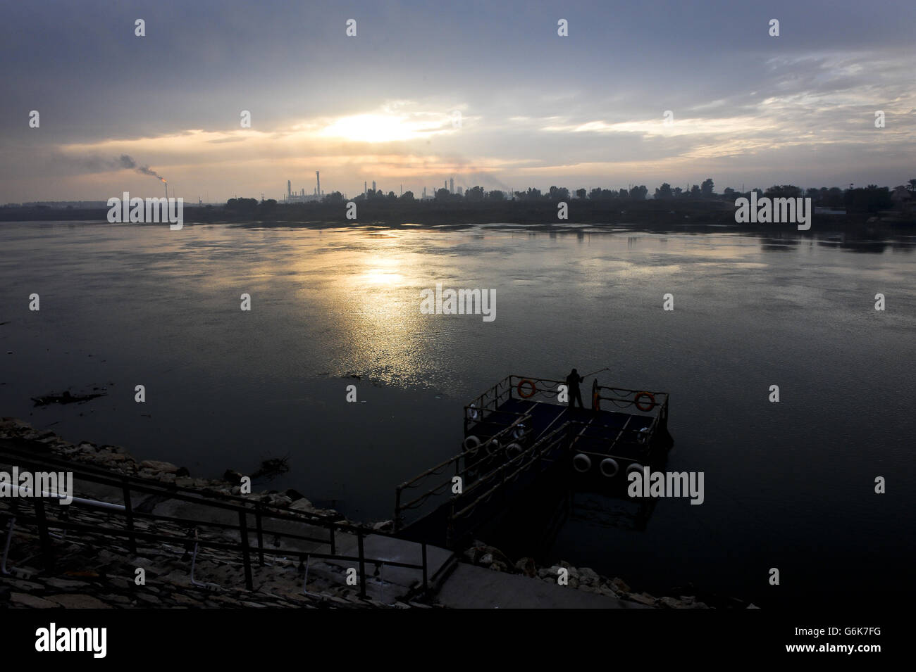 Ein Mann fischt bei Sonnenaufgang Müll aus dem Tigris River mit der Dora Ölraffinerie, im Hintergrund abgebildet, Bagdad. Stockfoto