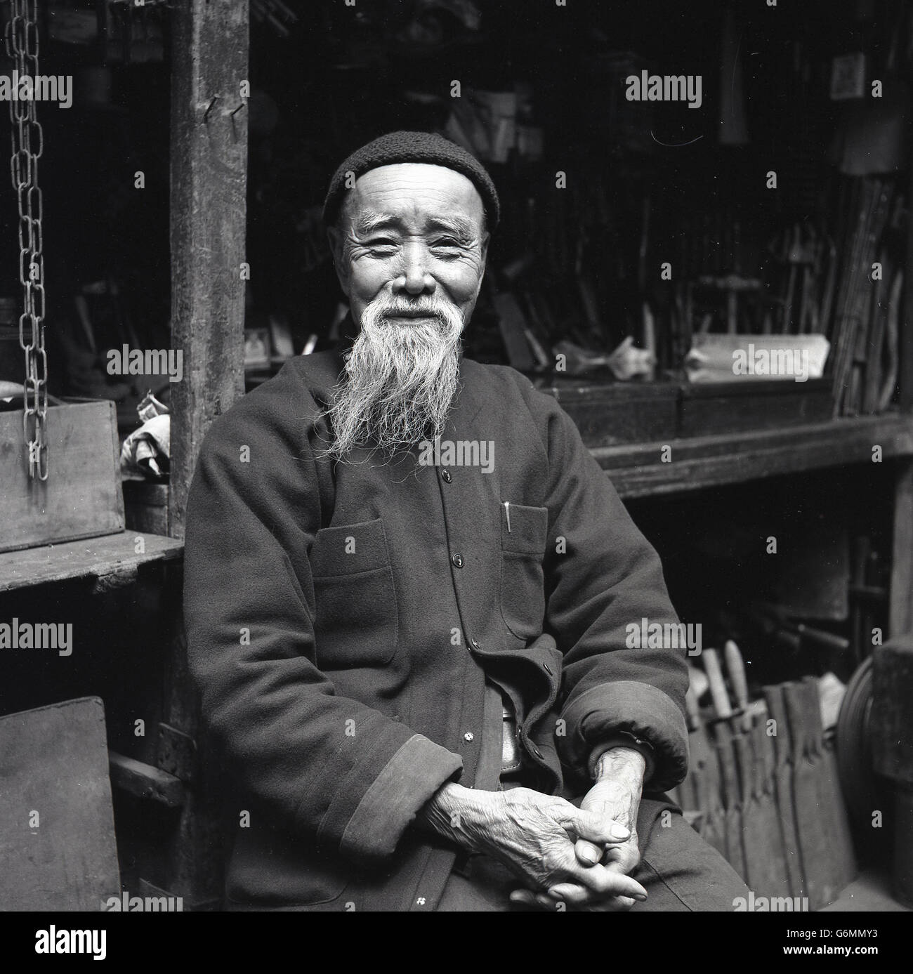 der 1950er Jahre, historische, Porträt des chinesischen Arbeiter sitzt stolz durch seine Werkzeuge tragen eine traditionelle Jacke und Mütze. Stockfoto