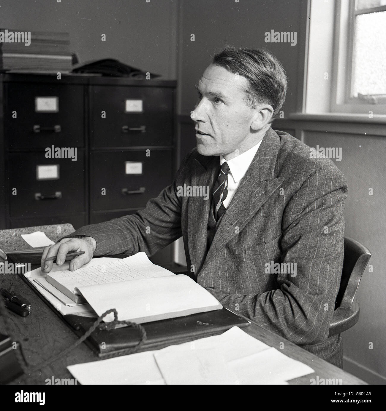 1950, historisch, ein leitender Angestellter in der britischen Industrie, der Anzug und Zeit trägt, in seinem Büro, das mit Papierkram, Tintenfleck und Metallablagen in der Ecke an seinem Schreibtisch sitzt. Stockfoto