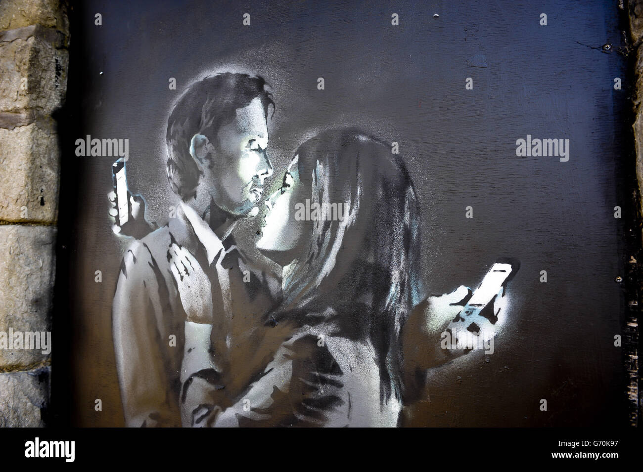 Ein Blick auf das neueste offiziell bestätigte Banksy Kunstwerk namens Mobile Lovers, mit einem Mann und einer Frau umarmt und Blick auf ihre Mobiltelefone, die auf eine schwarze Holzplatte in einem abgesperrten Eingang in Clement Street, Bristol, in Sichtweite des belebten Stadtzentrums gesprüht wird. Stockfoto