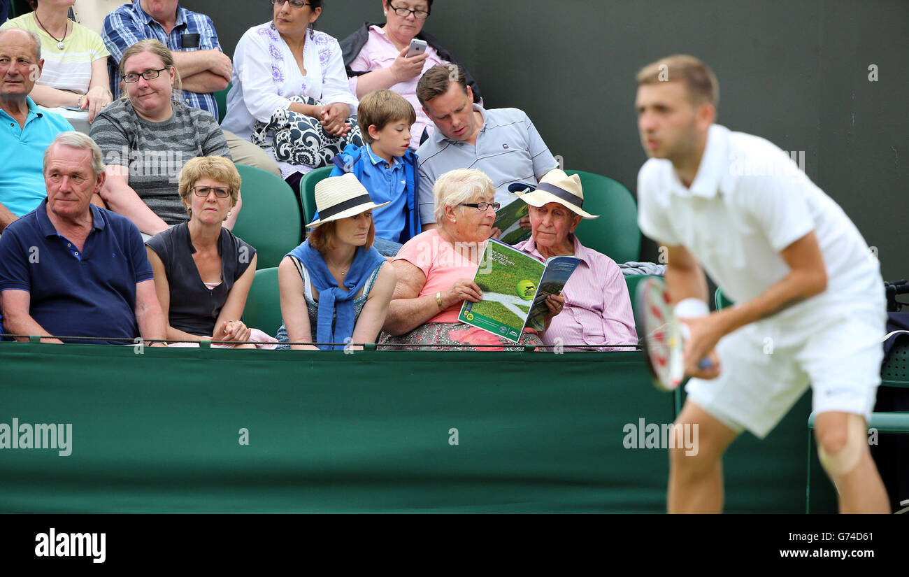 Ein Paar las ein Programm in der ersten Reihe, während der britische Daniel Evans (rechts) den russischen Andrey Kuznetsov während eines Tages bei den Wimbledon Championships im All England Lawn Tennis and Croquet Club, Wimbledon, antritt. Stockfoto