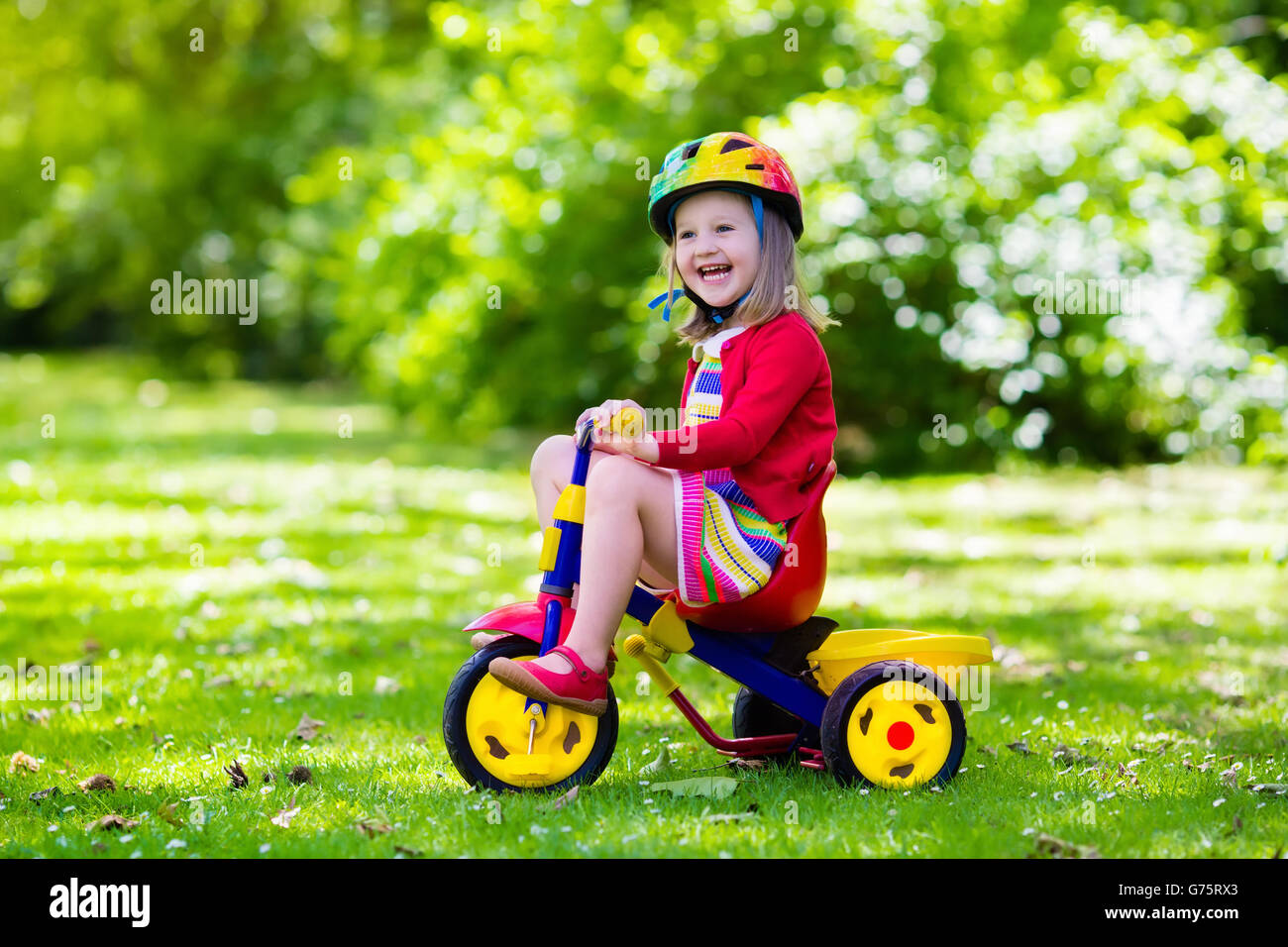 Niedliche Mädchen reiten ihr Dreirad im sonnigen Sommerpark-Schutzhelm tragen. Kinder Fahrrad. Erstes Fahrrad für kleines Kind. Stockfoto